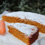 Slice of a carrot cake in the front and half of carrot cake on the behind with so powdered sugar above