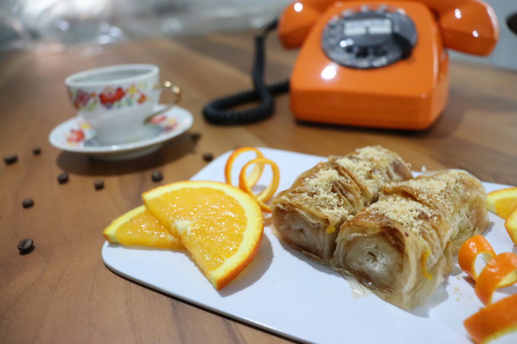 Zwei Baklava Stückchen auf einem weißen Teller dekorieret mit halbierten Orangenscheiben. Im Hintergrund ist ein organgenes, altes Telefon und eine nostalgische Tasse mit Blumenmuster zu sehen. 