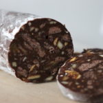 A piece of chocolate salami covered with powdered sugar. Inside with chocolate biscuits, nuts and dried fruits. In the front there are two slices of the chocolate salami.